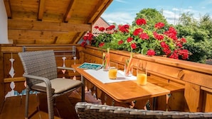 Eine Holzterrasse im Freien mit Tisch und Stühlen, umgeben von roten Blüten und Grünpflanzen.