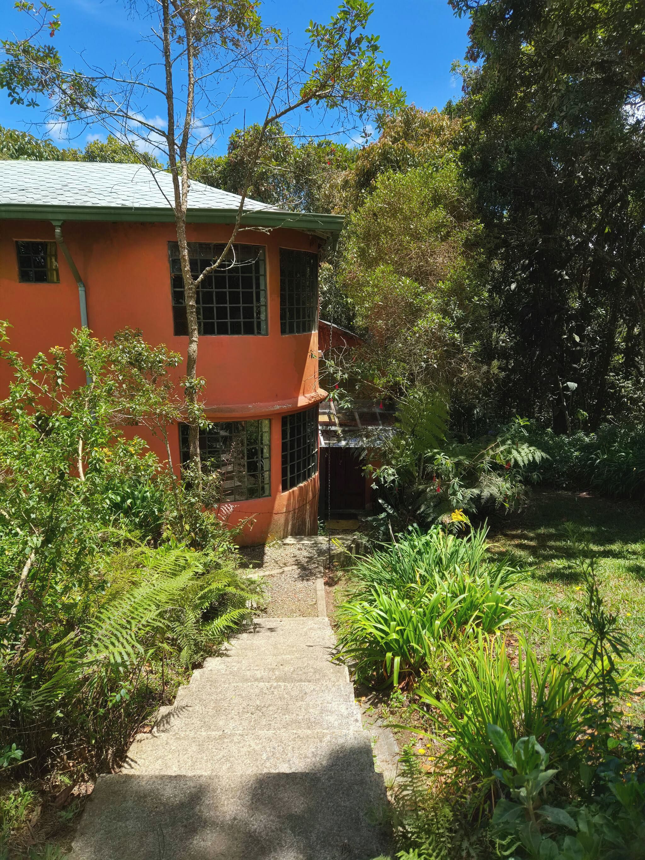 A building with a curved roof, surrounded by dense greenery and a gravel pathway leading to it.