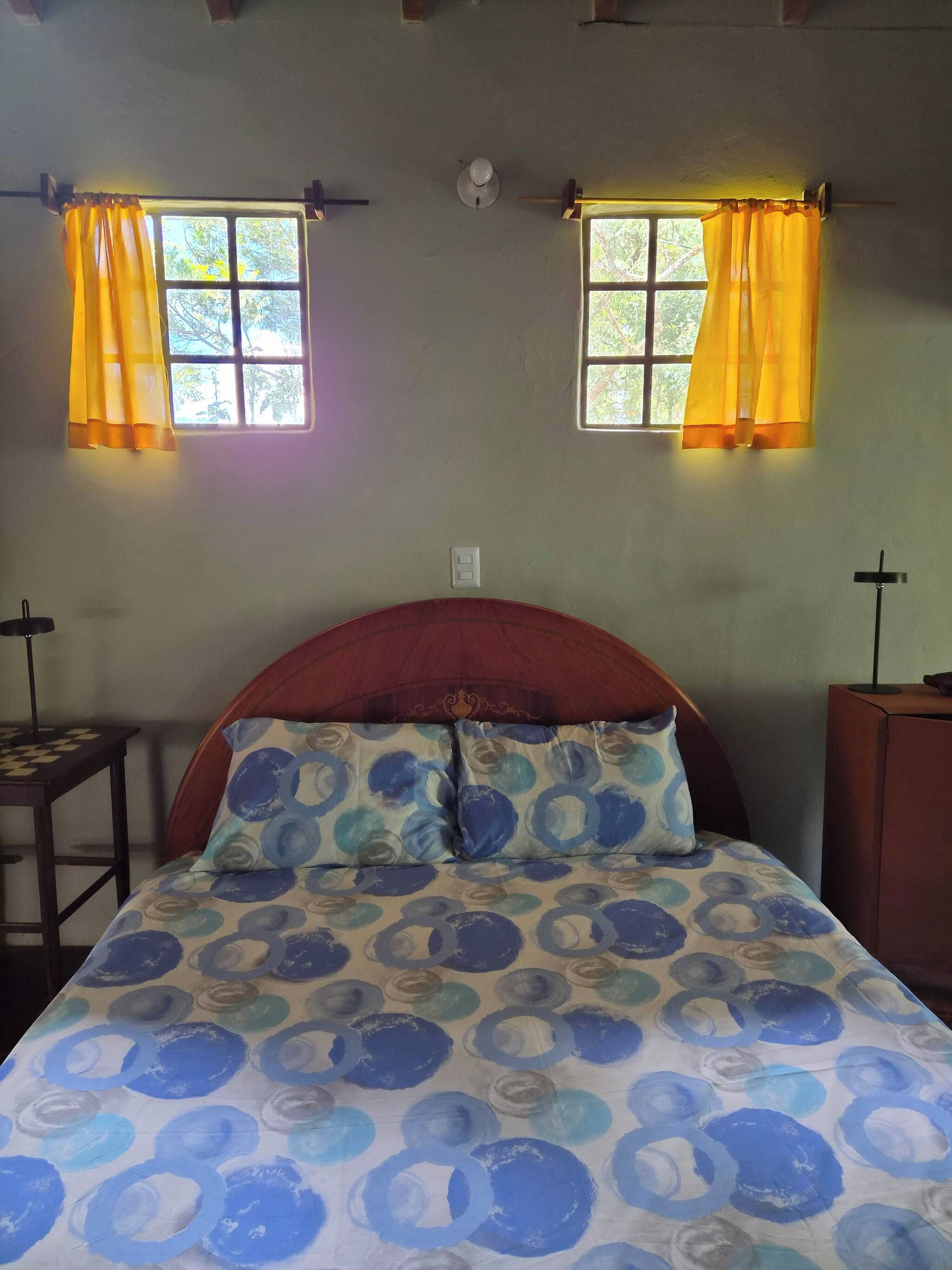 A room with a bed, two windows with curtains, a bedside table, and a cross on the wall.