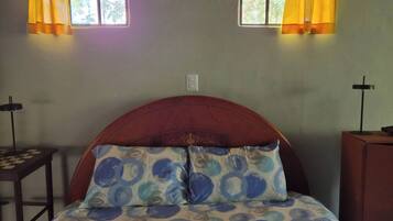 A room with a bed, two windows with curtains, a bedside table, and a cross on the wall.