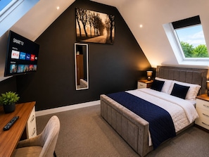 Een slaapkamer met een schuine balkenlaag, een aan de muur bevestigde televisie, een bed met hoofdbord, een nachtkastje met lamp, een bureau met een plant en een raam met uitzicht op bomen.