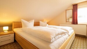 Een eenpersoonsbed met witte lakens in een kamer met een patroon vloer, een nachtkastje met een lamp en een raam met gordijnen.
