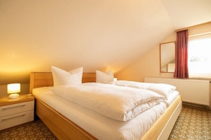 Uma cama de solteiro com roupa de cama branca em quarto com piso estampado, mesa de cabeceira com abajur e janela com cortinas.