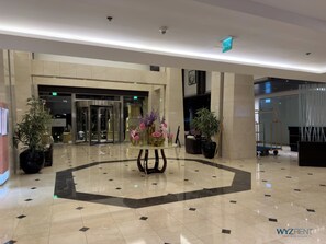 Un vestíbulo de hotel con una mesa redonda central, plantas en macetas y una entrada de vidrio.