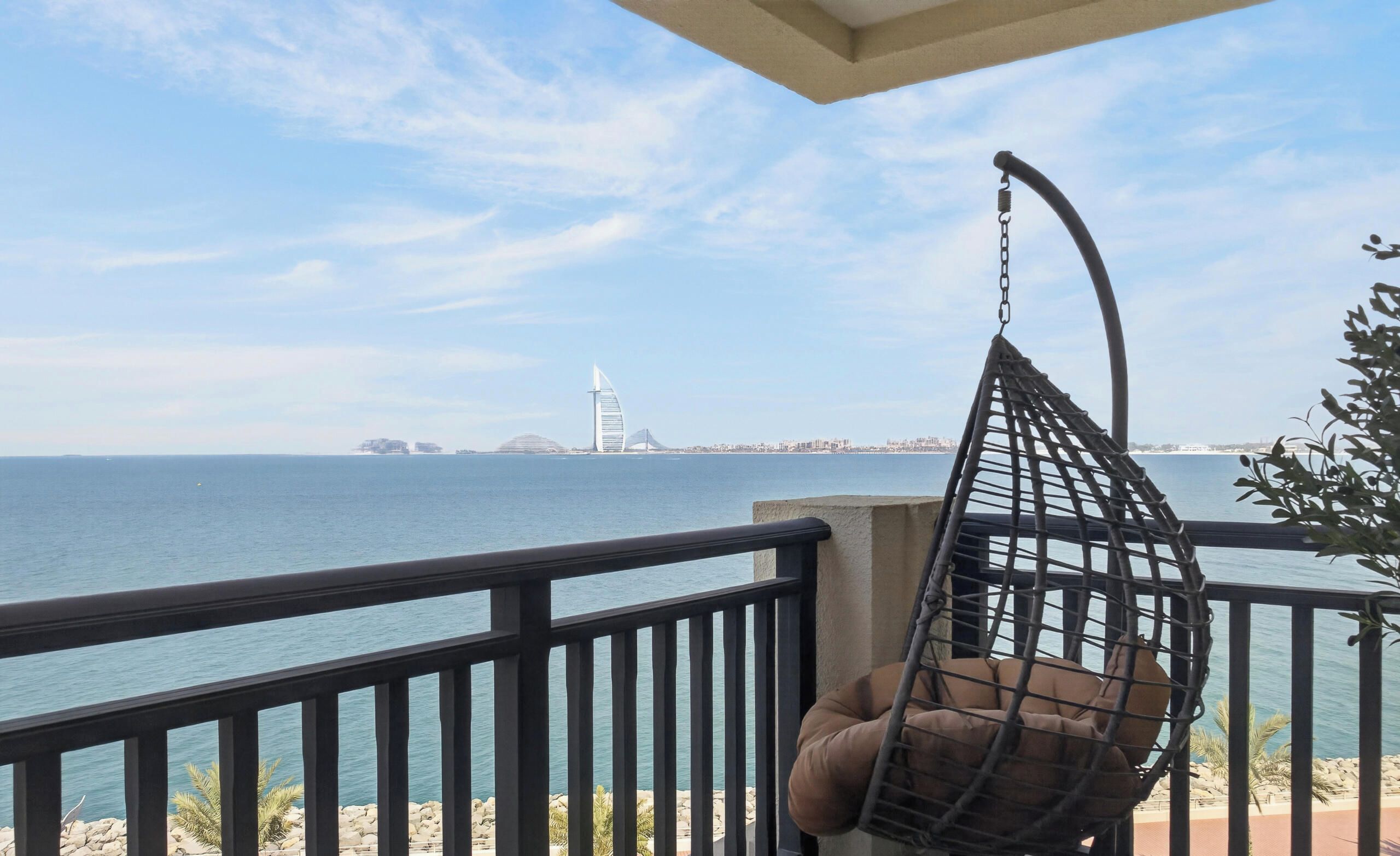A balcony with a hanging chair overlooking a body of water, a distant city skyline, and a clear sky.