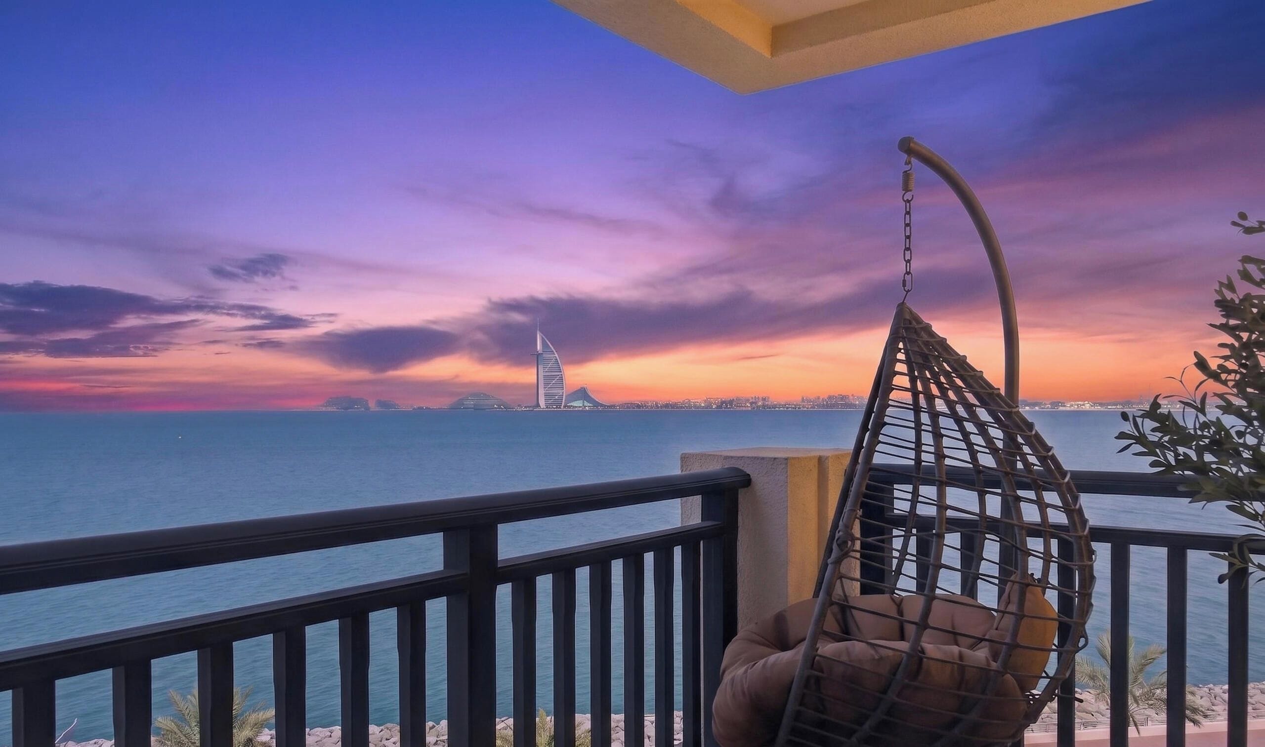 A balcony with a view of a city skyline at sunset, a hanging chair, and a railing.