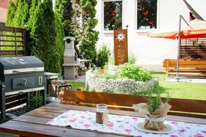 Eine Terrasse im Freien mit einem Holztisch, einem Grill, einem Steinpflanzgefäß mit Grünpflanzen und einer Holzbank.