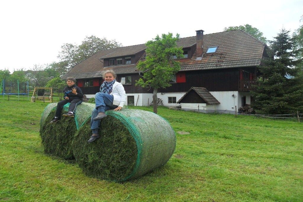 A rural setting with a house, hay bales, and a person sitting on a hay bale.