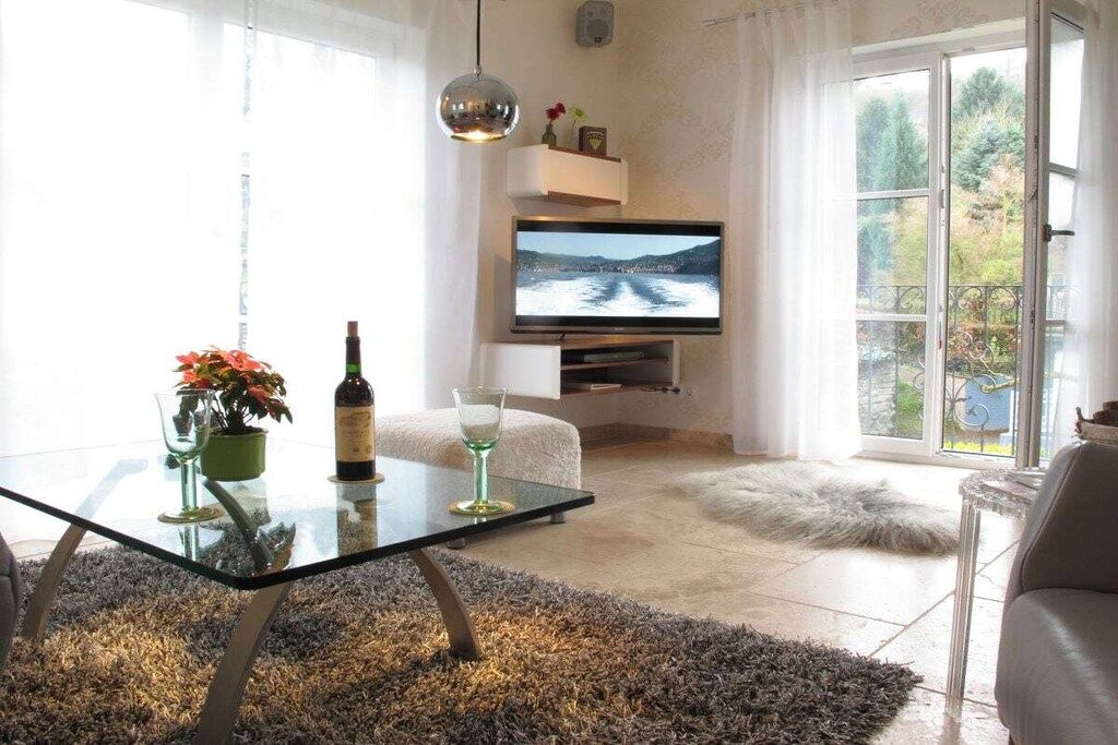 A living room with a glass coffee table, a sofa, a television mounted on the wall, and a view of a lake through the window.