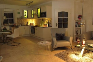 A modern kitchen with white cabinets and a dining area with a white chair. There is a glass table with a candle and a bottle. - Holiday home in Remagen with swimming pool (Remagen)