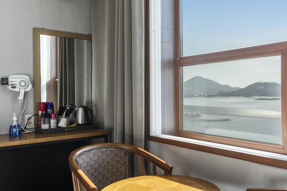 Habitación de hotel con un ventanal grande que ofrece vistas a un cuerpo de agua y montañas, una silla de madera, una mesita con artículos de aseo y un secador de pelo en la pared.