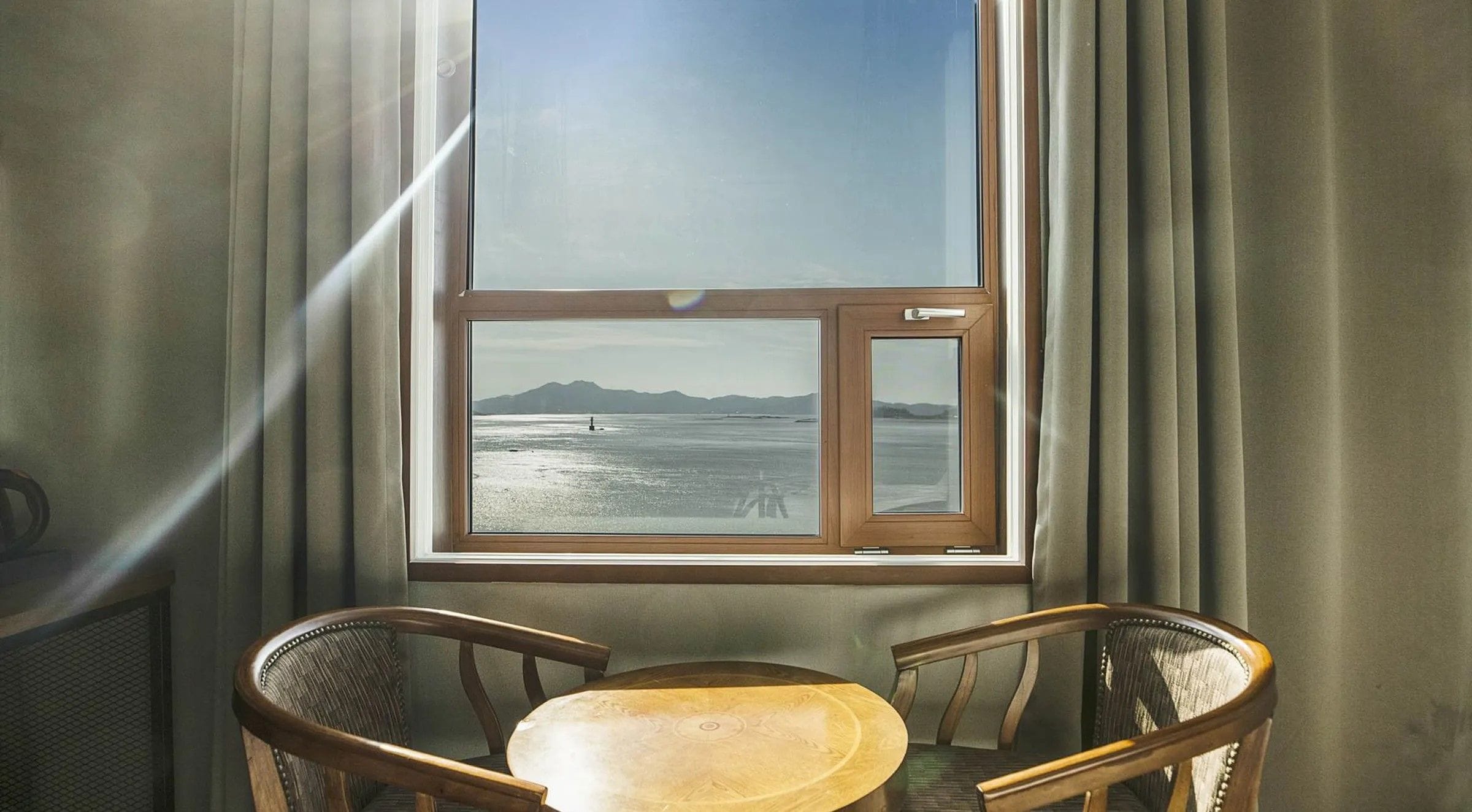 A room with a large window overlooking a body of water and distant mountains. There are two wooden chairs and a small table in the foreground.