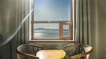 A room with a large window overlooking a body of water and distant mountains. There are two wooden chairs and a small table in the foreground.