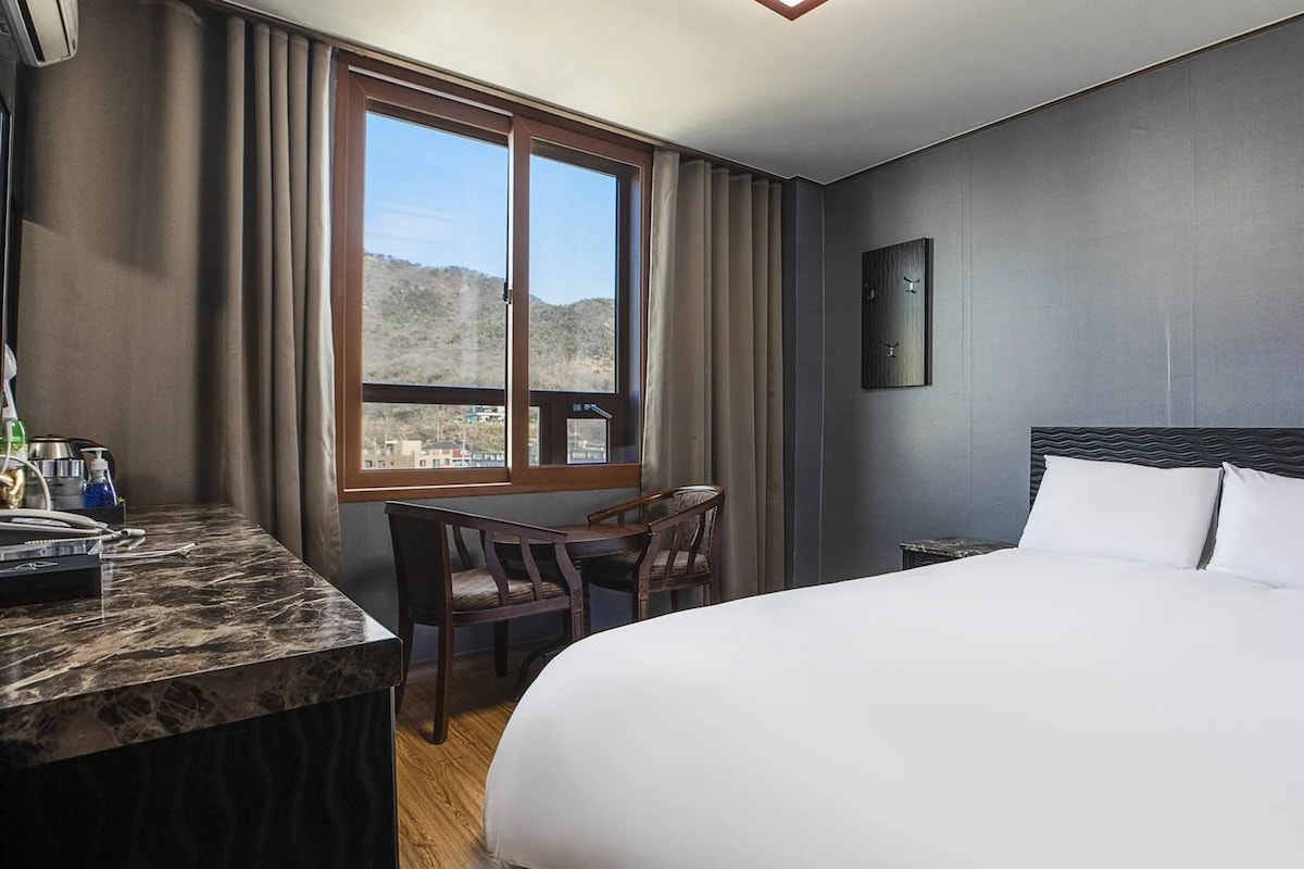 Habitación de hotel con cama, escritorio con silla, ventana con cortinas e imagen colgada en la pared.