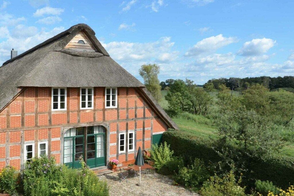 Une maison traditionnelle en briques, avec un toit de chaume, une porte verte et un petit jardin agrémenté d’un banc et de plantes en pot.