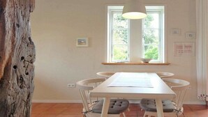 Un área de comedor con una mesa y sillas blancas, una lámpara colgante, un ventanal con vistas a la vegetación y un tronco de árbol de madera.