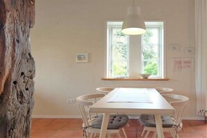 A dining area with a white table and chairs, a hanging light fixture, a window with a view of greenery, and a wooden tree trunk. - "On the dike" - holiday apartment (Bremen)