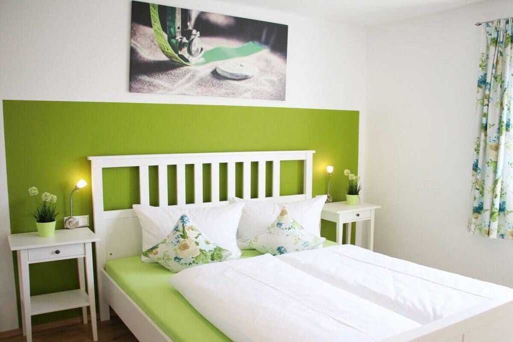 A bed with white bedding, green pillows, and a white headboard. There are two bedside tables with lamps and a small plant on each. A painting is visible on the wall above the bed.