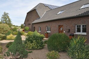 Un edificio de ladrillo con techo inclinado, paneles solares y un jardín bien cuidado con diversos arbustos y un conífero.