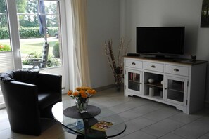 A living room with a black leather chair, a glass coffee table with a vase of flowers, a white cabinet with a television, and a view of the outdoors through a glass door. - At the Steenbrukshof woods (Xanten)