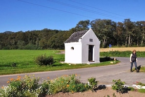 A small white building with a pitched roof, located beside a road with a clear sky and green fields in the background. - At the Steenbrukshof woods (Xanten)