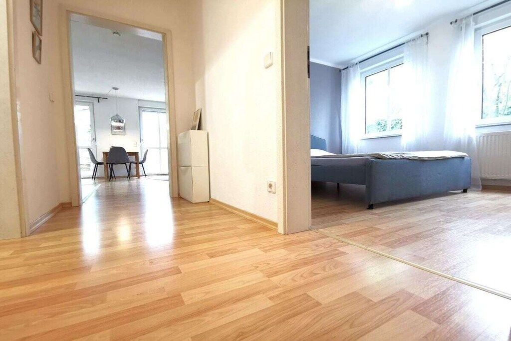 A room with a wooden floor, a bed, a window, and a door leading to another room.