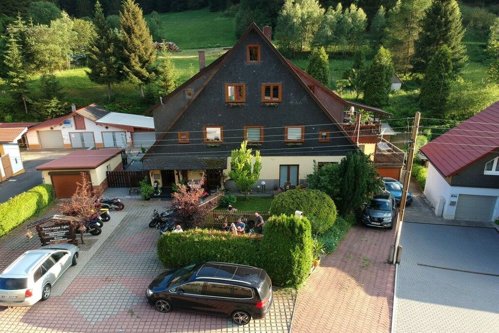 A large two-story house with a sloped roof, surrounded by a paved driveway and parked cars.