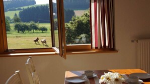 Une pièce avec une table à manger dressée pour deux personnes, une fenêtre avec des rideaux et une vue sur des chevaux dans un champ.
