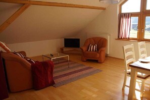 A cozy loft-style room with a wooden ceiling, a sofa, a chair, a small table, and a window with curtains. - Panoramic view of the Höllbach valley (Rettenbach)