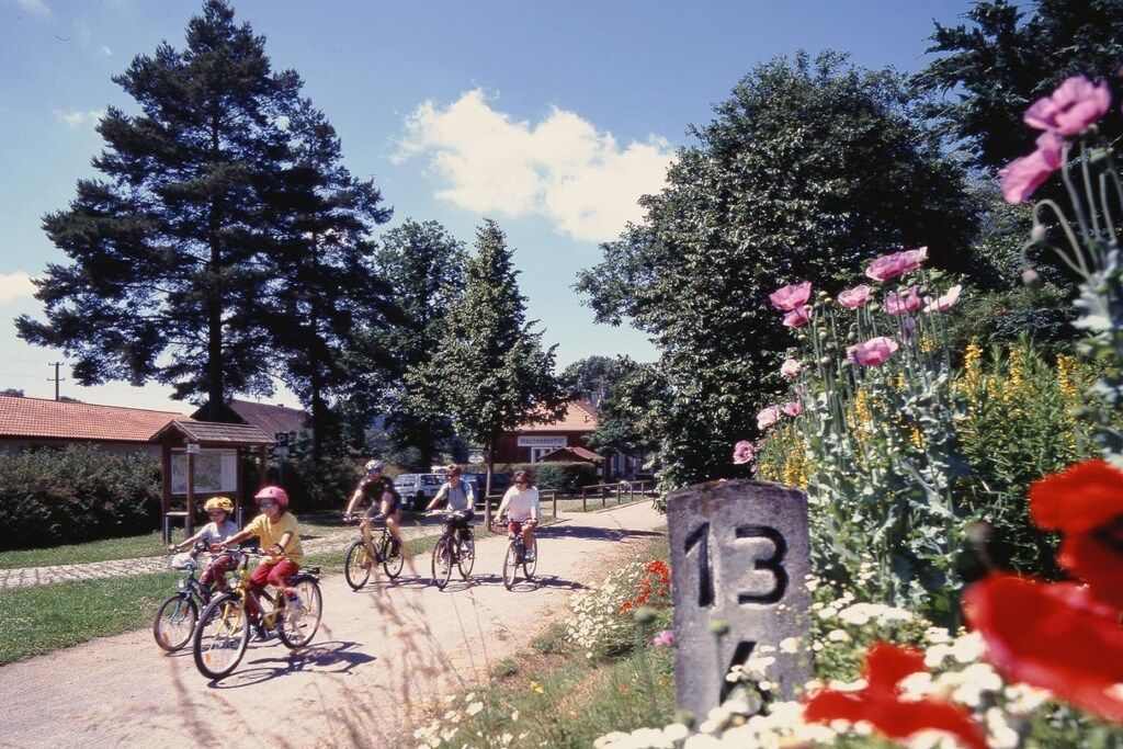 A paved path with cyclists, surrounded by greenery and a building with a red roof.