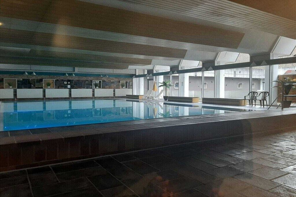 An indoor swimming pool with clear water, surrounded by a tiled floor and walls, and a ceiling with a geometric pattern.