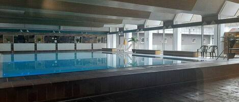 An indoor swimming pool with clear water, surrounded by a tiled floor and walls, and a ceiling with a geometric pattern.