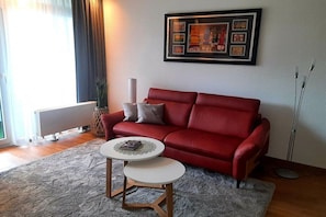 Ein Wohnzimmer mit einem roten Sofa, zwei kleinen weißen Tischen, einem gerahmten Kunstwerk an der Wand und einer Heizung.