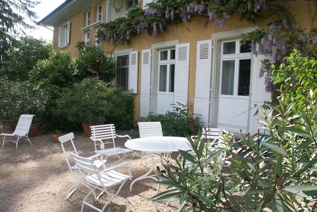 Ein Gebäude mit einem Garten und einer Terrassenanlage, ausgestattet mit weißen Metallstühlen und einem Tisch, umgeben von üppiger Bepflanzung und blühenden violetten Weinranken.