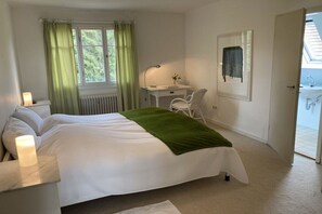 A bedroom with a bed, a desk, a chair, a radiator, a window with curtains, and a door leading to a bathroom. - 1 Alpenblick - Steineckhof (Schopfheim)