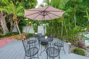 Un pati exterior amb una taula rodona i cadires, un parasol gran i vegetació tropical exuberant.