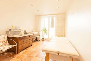 A room with a bed, a wooden dresser, a desk, a chair, a plant, and a window with blinds.