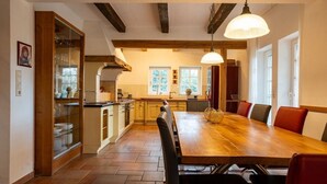 A spacious kitchen with a large wooden table and red chairs, a hanging light fixture, and a view of the outdoors through the windows.