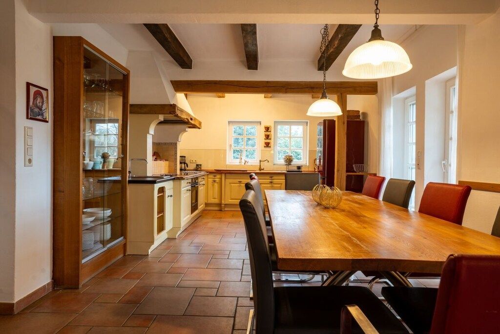 Eine geräumige Küche mit einem großen Holztisch und roten Stühlen, einer Hängelampe sowie Blick ins Freie durch die Fenster.