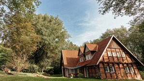A traditional half-timbered house with a sloped roof, surrounded by a well-maintained lawn and dense trees.