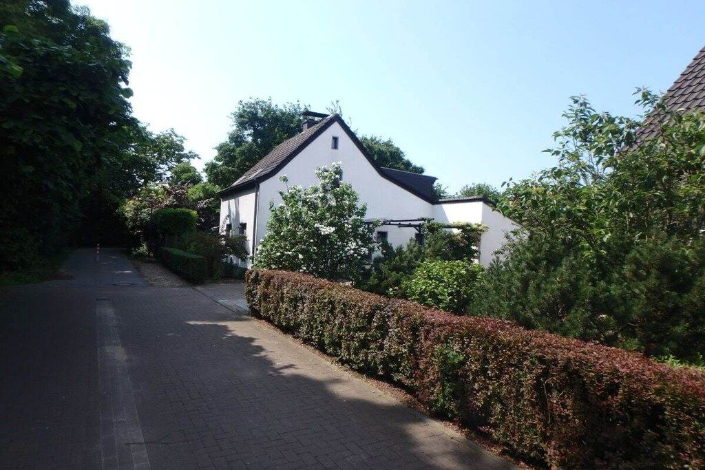 A residential street with a white house, a paved road, and a hedge.