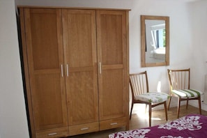 Ein Schlafzimmer mit großem Holzschrank, zwei Stühlen, einem Bett mit gemustertem Überwurf und einem Spiegel an der Wand.