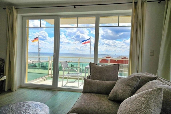 Une pièce comprenant un canapé, un pouf rond et offrant une vue sur la mer grâce à de grandes fenêtres.