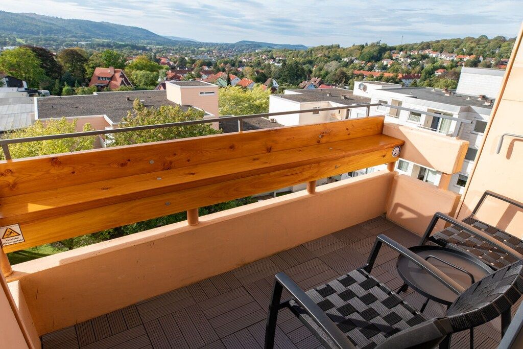 A balcony with a wooden bench and two chairs, overlooking a residential area with houses and greenery.
