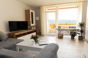 A modern living room with a flat-screen TV, grey sofas, a white coffee table, and a balcony with a view. - Holiday apartment with 1 bedroom (Bad Harzburg)