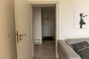 A room with a door leading to a hallway, a small white bench, and a grey sofa with a pillow. - Holiday apartment with 1 bedroom (Bad Harzburg)