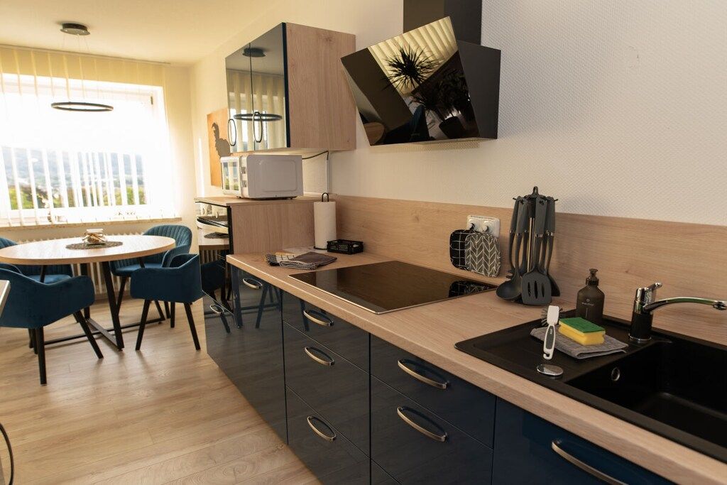 A modern kitchen with a central island, a dining area with blue chairs, and a large window providing natural light.