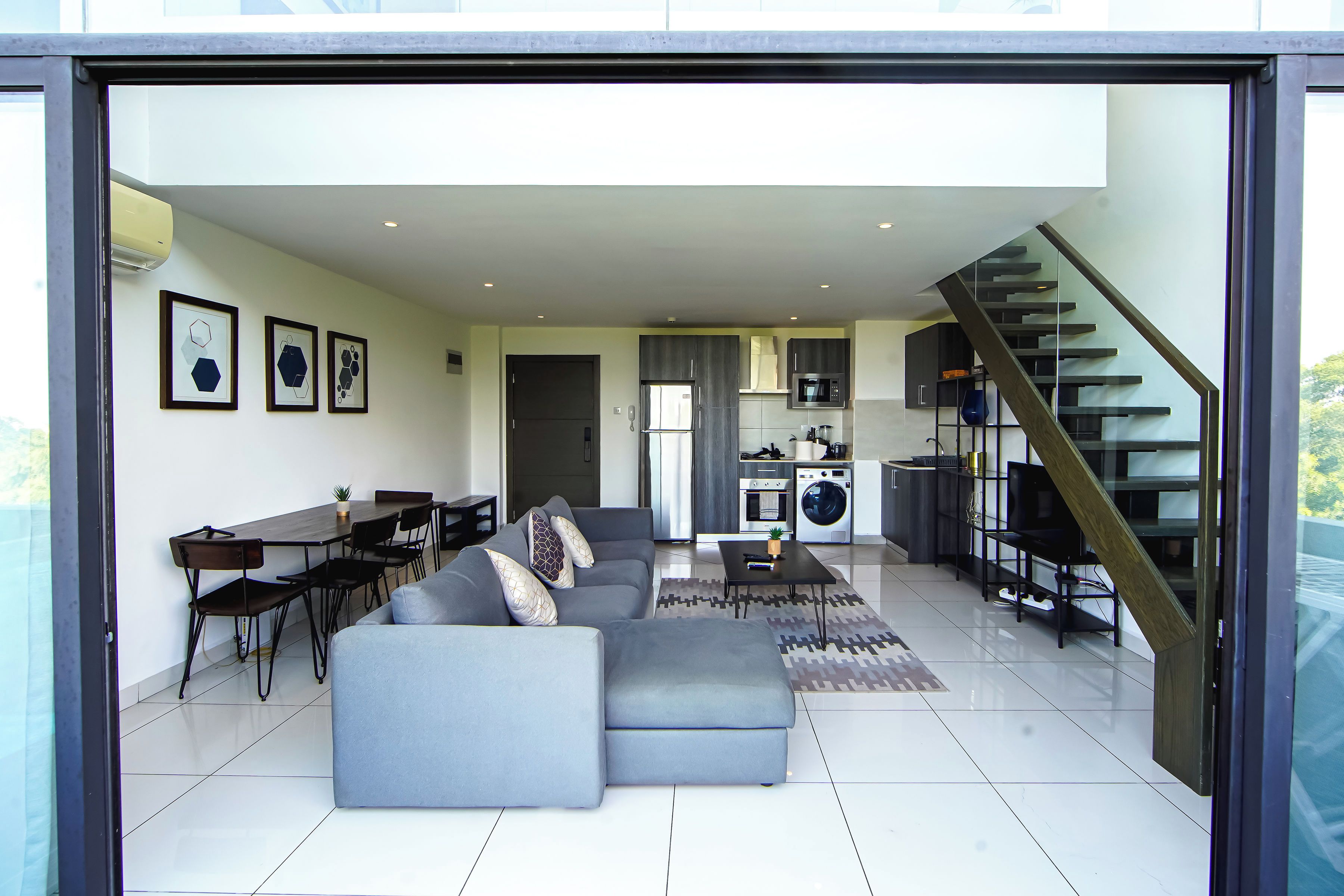A modern living space with a dining area, a sofa, a staircase, and a kitchen.