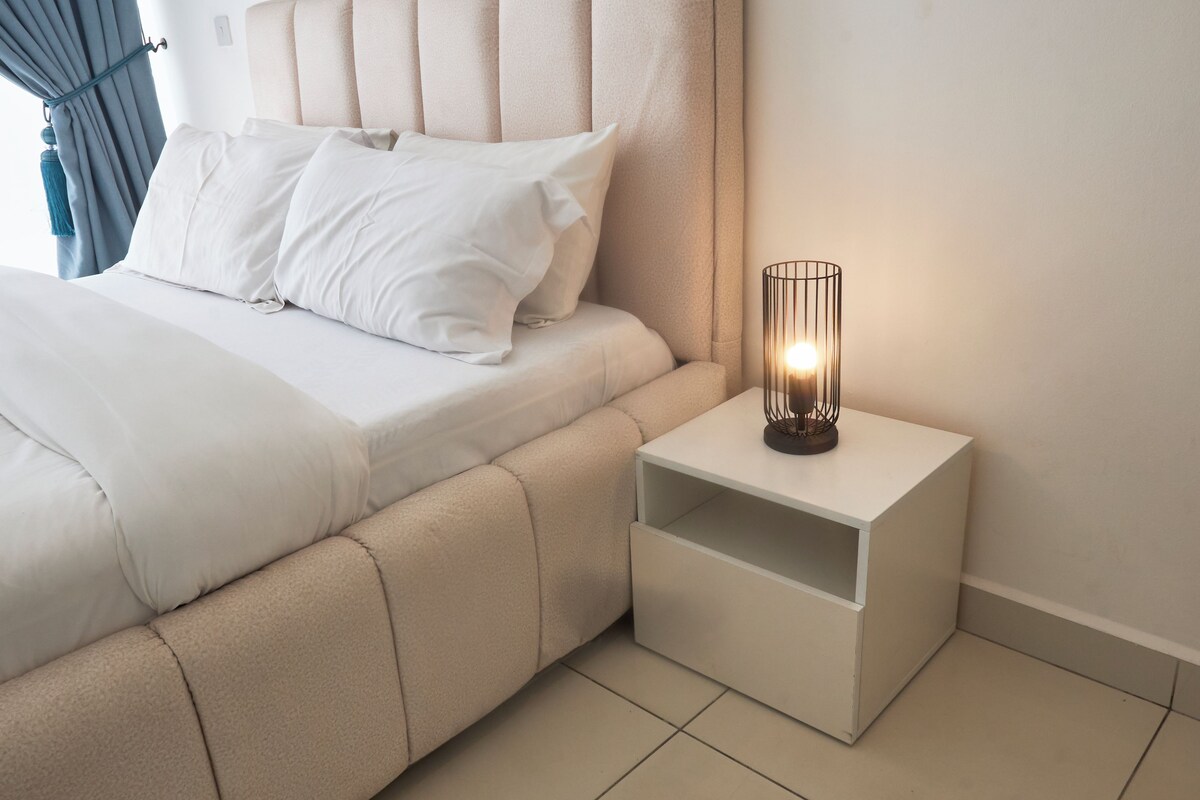 Ein ordentlich gemachtes Bett mit weißen Leinen und Kissen, ein Nachttisch mit Lampe sowie ein Blick auf den gefliesten Boden des Zimmers.