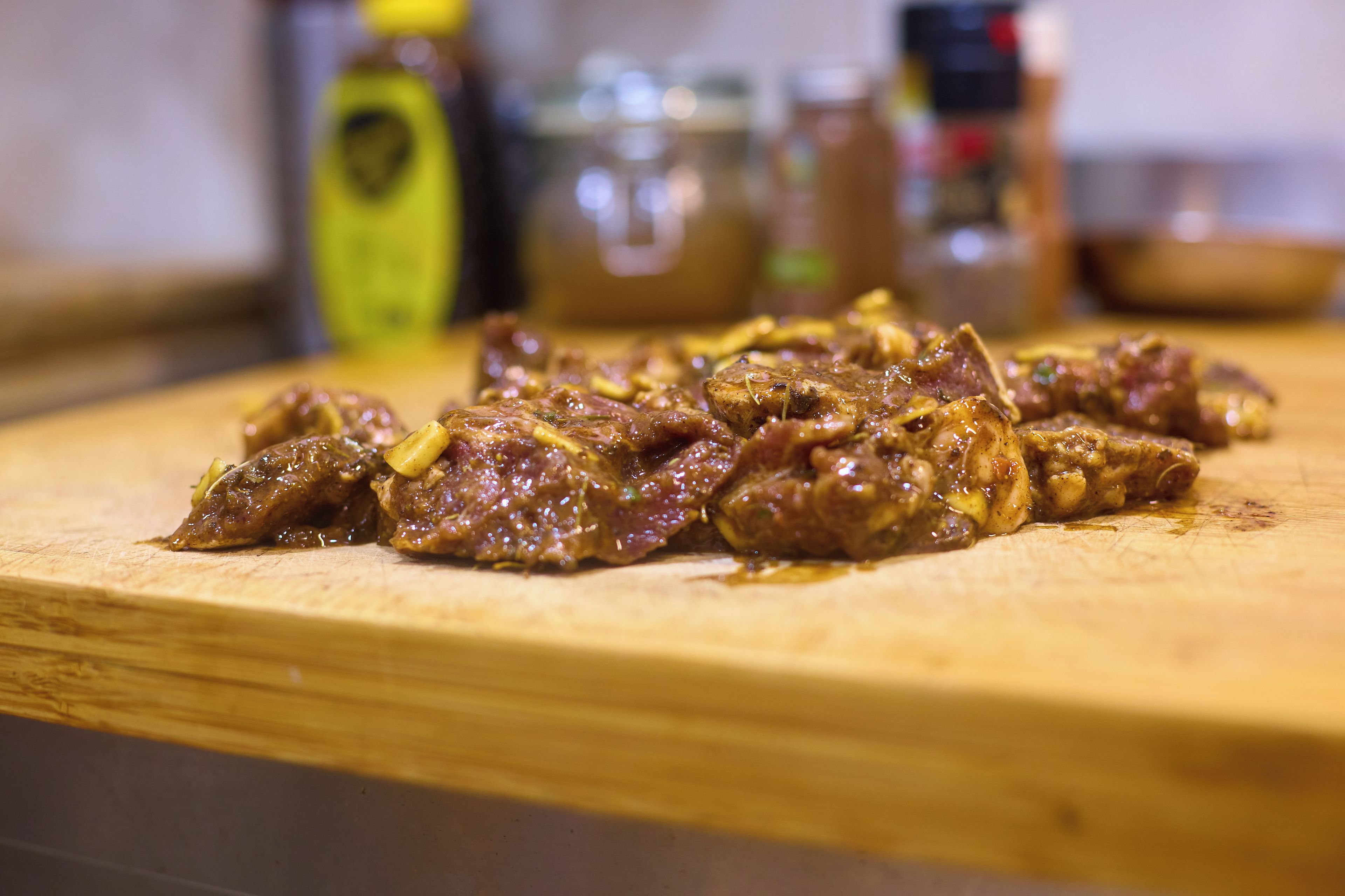 A wooden cutting board with a dish of cooked meat, possibly beef, in a sauce, with blurred kitchen items in the background.